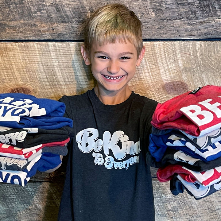 Be Kind to Everyone® Youth Apparel Collection image featuring Hudson holding a variety of shirts from the shop.