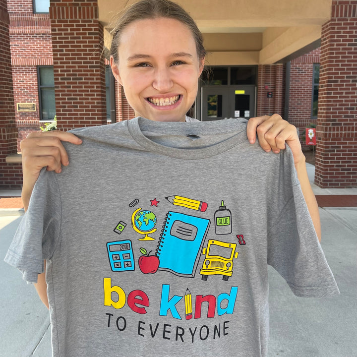 Jordyn, displaying our NEW School Be Kind to Everyone® short-sleeved tee in front of a school.