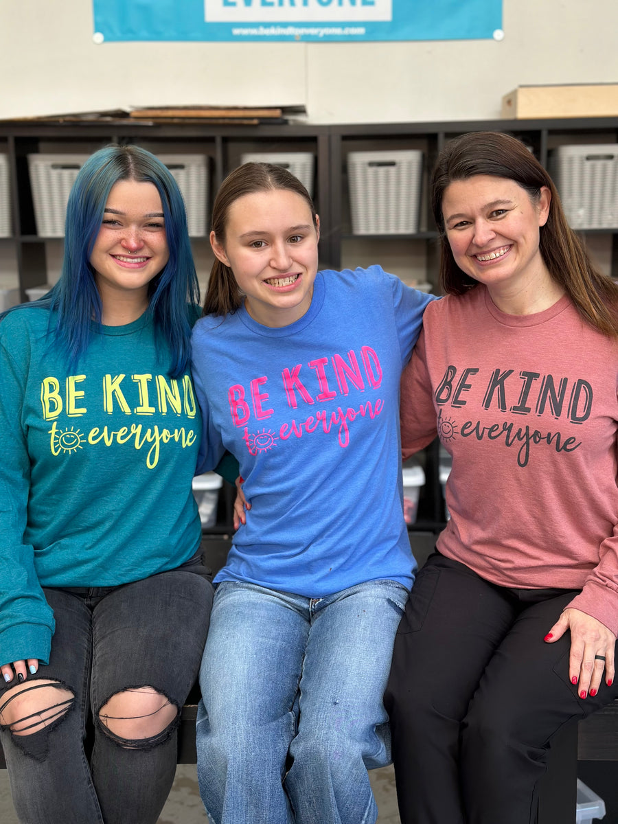 Our Long Sleeve Surprise Packs feature our Smiling Sun Be Kind to Everyone® design on a heathered mauve, heathered royal, or heathered teal long-sleeved tee.  Jordyn is wearing a small, and Jackie and Sarah are wearing mediums in this picture. 