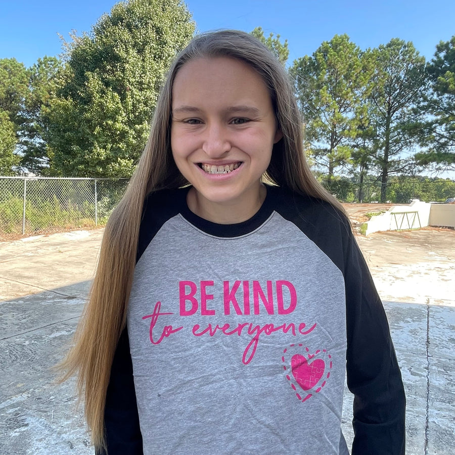 Our Original Jordyn Be Kind to Everyone® Raglan tee is a heather gray with heather black sleeves and a bright pink design.