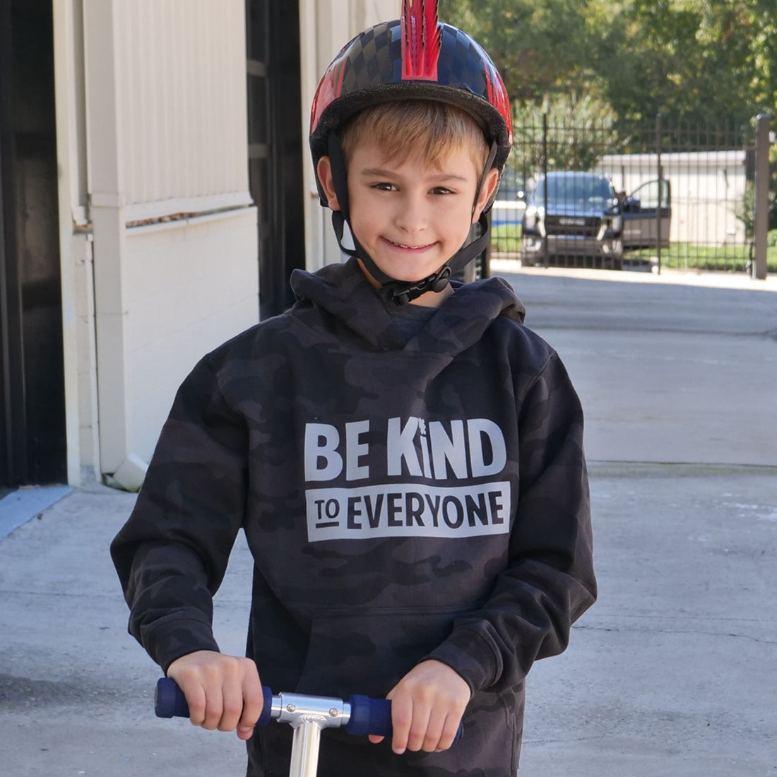 Hudson, on his scooter outside the shirt shop, in a Hudson Be Kind to Everyone® black and gray camo youth hoodie. 